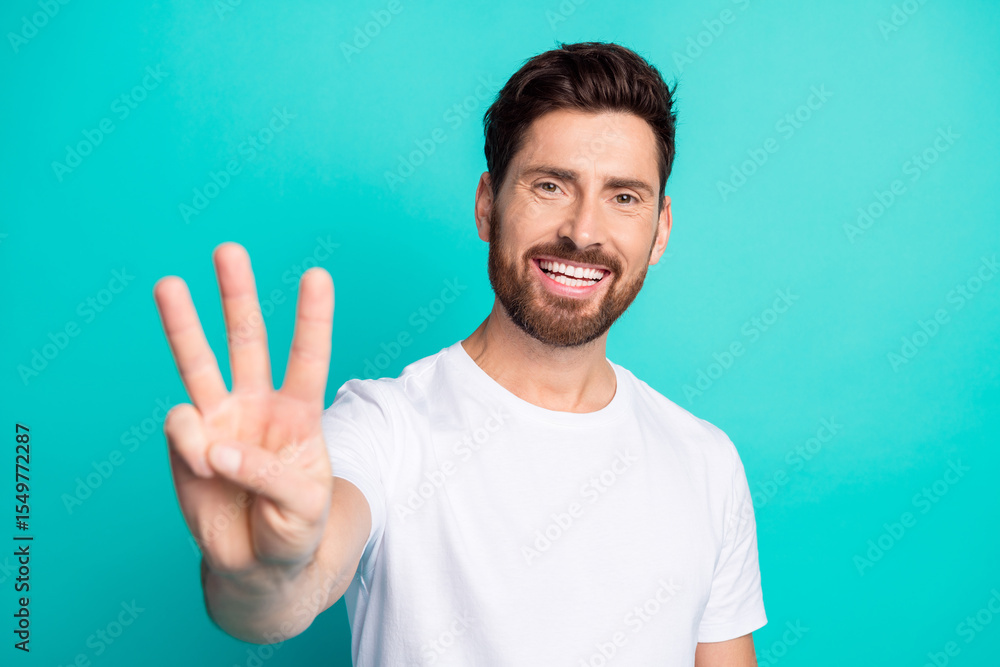 Naklejka premium Smiling young man showing three fingers in casual white t-shirt with turquoise background as a sign of positivity