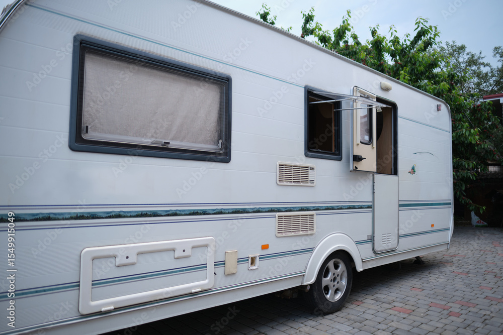 Naklejka premium A white camper trailer is parked on a paved backyard near a brick house and cherry tree. RV travel content, camping promotions, suburban lifestyle visuals, or articles about alternative housing