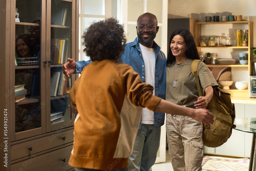 Fototapeta premium Caucasian woman and Black man standing together smiling while welcoming curly haired child running toward them in home setting, military dog tag visible on woman, family reunion scene