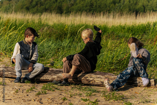 Bilde på lerret Three children sit and play on a driftwood log at the edge of a grassy dune, enjoying a sunny and breezy day in nature