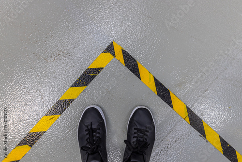 A person's feet in black shoes stand on a grey concrete floor, bordered by a prominent yellow and black striped hazard warning line.
