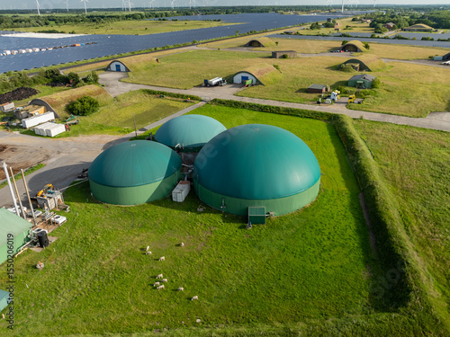 Aerial view of a renewable energy site with biogas domes, sheep grazing, solar field and repurposed munitions depot, showcasing integrated sustainable infrastructure in rural landscape.