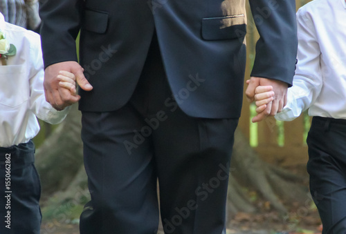 Closeup view of father and kids holding hands during wedding ceremony wearing formal clothing while walking down the aisle.