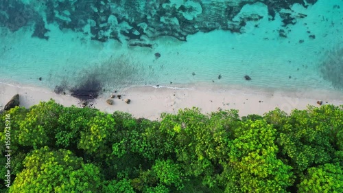 4K birdseye drone footage of Nusa Dua coastline in Bali. Blue ocean waves crash on tropical beach with lush green forest along shoreline. Bright, tropical, commercial-style shot.
