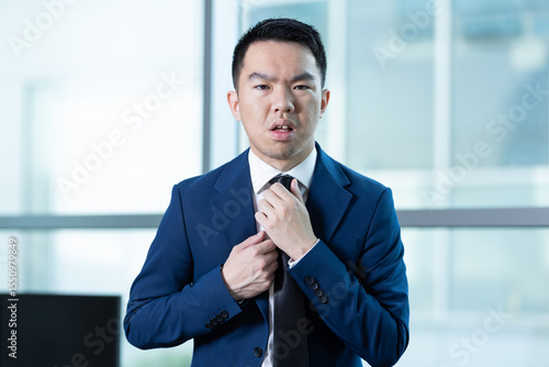 A young man in a suit adjusts his tie with a tense expression. The photo conveys a feeling of impostor syndrome, anxiety, or performance pressure in a corporate environment.