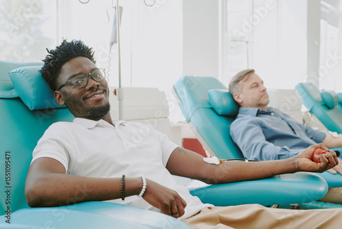 Foto Young Black man donating blood while sitting in medical chair, smiling at camera