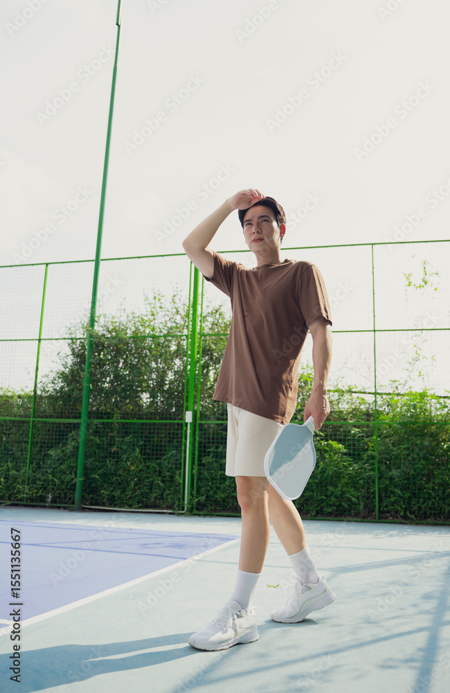Fototapeta premium Young Asian man enjoying a relaxing moment on a pickleball court in bright afternoon light