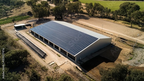 Aerial view of a large agricultural building with extensive solar panel installation on its roof, surrounded by greenery and infrastructure