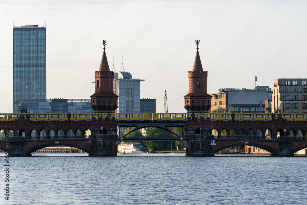 Fototapeta premium Wide view of Oberbaumbrucke in Berlin with a yellow U-Bahn train crossing above the river. The twin brick towers and modern skyline highlight the city s architectural contrast.