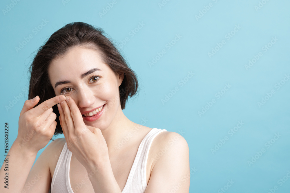 Fototapeta premium Woman putting in contact lens on light blue background. Space for text