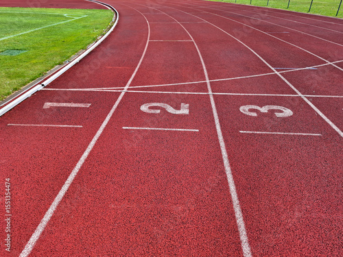 Fotografie Curving red synthetic running track with lanes 1, 2, and 3, stark white markings and numbers, flanked by lush green grass