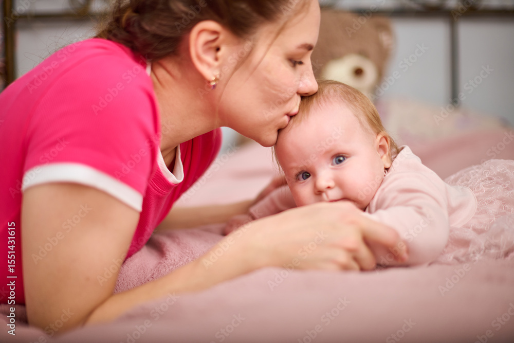 Young mother with braided hair kisses light-haired baby girl on pink bedspread, expressing warmth and love. Soft lighting in cozy bedroom setting enhances tenderness Young mother with braided hair kisses light-haired baby girl on pink bedspread, expressing warmth and love. Soft lighting in cozy bedroom setting enhances tenderness