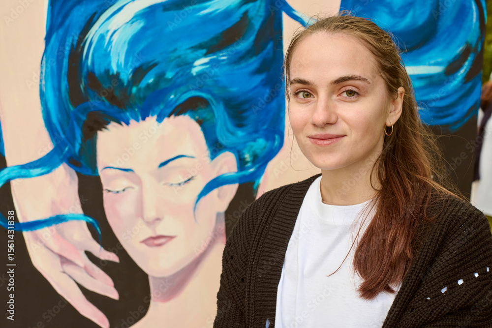 Fototapeta premium Young female artist with brown hair stands beside her vibrant painting. Artwork, depicting serene face with flowing blue hair, contrasts with her calm smile