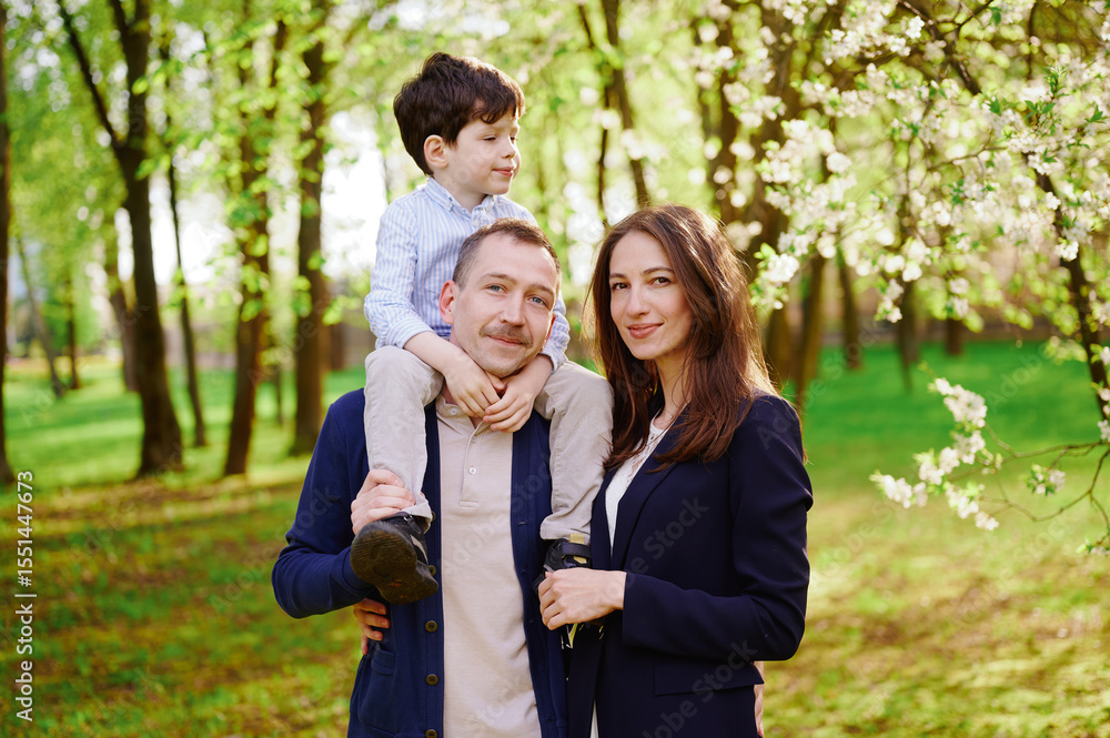happy family enjoying spring day outdoors. mother, father with child on shoulders in sunny park scenery. blossoming trees, smiling faces, joyful family expression, light jackets. happy family enjoying spring day outdoors. mother, father with child on shoulders in sunny park scenery. blossoming trees, smiling faces, joyful family expression, light jackets.