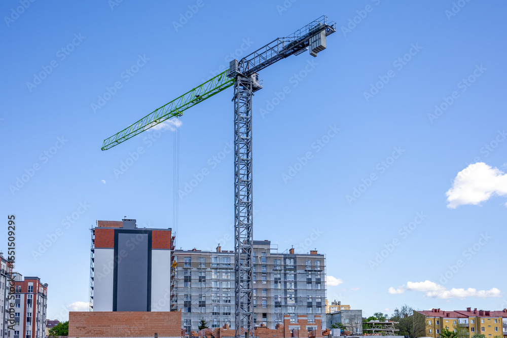 Tall tower crane operating on residential construction site with apartment buildings in background under clear blue sky
Tall tower crane operating on residential construction site with apartment buildings in background under clear blue sky