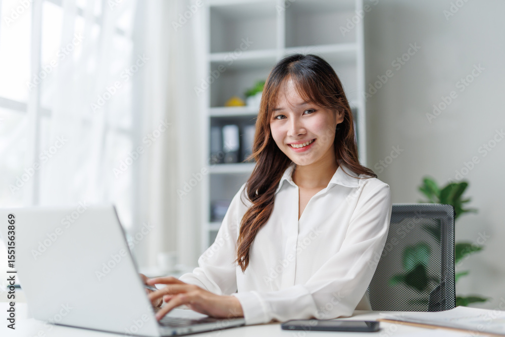 Smiling businesswoman sitting at a desk, using a laptop in a bright, modern office filled with natural light and a vibrant atmosphere Smiling businesswoman sitting at a desk, using a laptop in a bright, modern office filled with natural light and a vibrant atmosphere