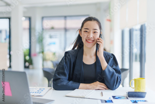 Young manager is speaking on smartphone and smiling while analyzing financial charts at her workplace