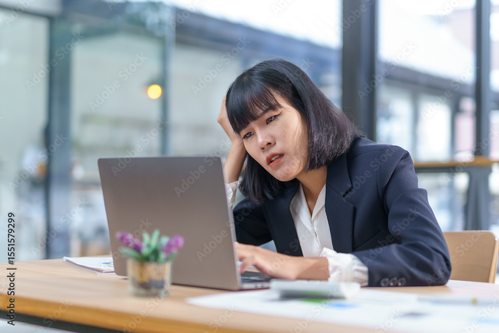 Worried Asian manager sitting at a desk in a modern office, using a laptop while facing challenges and pressures of corporate life Worried Asian manager sitting at a desk in a modern office, using a laptop while facing challenges and pressures of corporate life