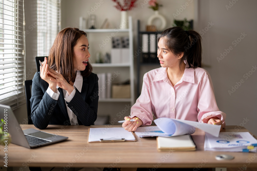 Two women are sitting at a desk, one of them is holding a piece of paper Two women are sitting at a desk, one of them is holding a piece of paper