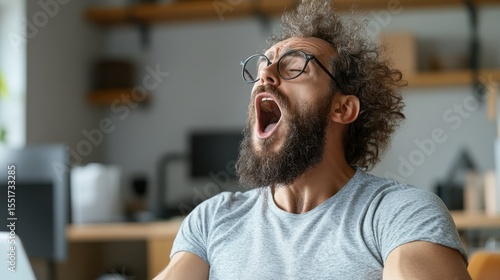 A young man with curly hair is yawning widely in an office environment, expressing fatigue and the common challenge of maintaining energy during work hours.