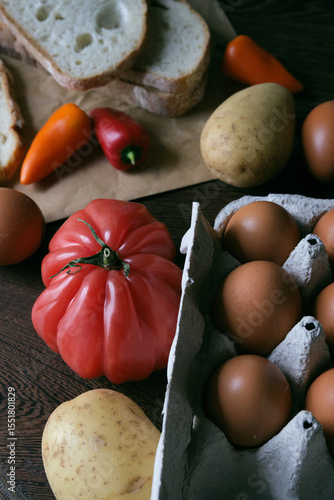 A rustic flat lay featuring slices of homemade artisan bread, fresh brown eggs in a carton, heirloom tomato, sweet peppers, and potatoes on a wooden kitchen table. Natural light and cozy.
