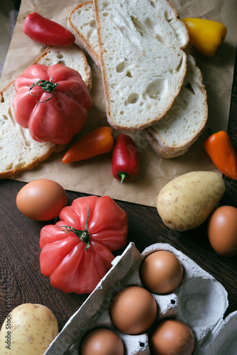 A rustic flat lay featuring slices of homemade artisan bread, fresh brown eggs in a carton, heirloom tomato, sweet peppers, and potatoes on a wooden kitchen table. Natural light and cozy.