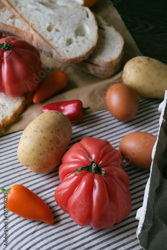 A rustic flat lay featuring slices of homemade artisan bread, fresh brown eggs in a carton, heirloom tomato, sweet peppers, and potatoes on a wooden kitchen table. Natural light and cozy.