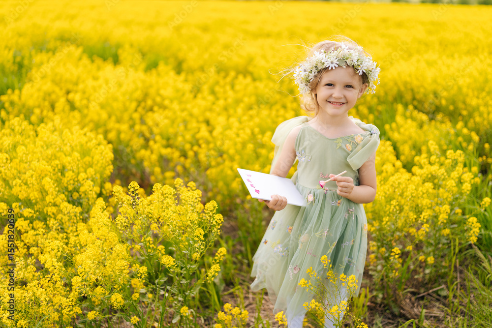 Fototapeta premium Cheerful child girl wearing flower wreath and fairy costume standing posing at blooming field during sunny summer day, drawing on canvas, enjoying beautiful nature. Concept of happiness childhood.