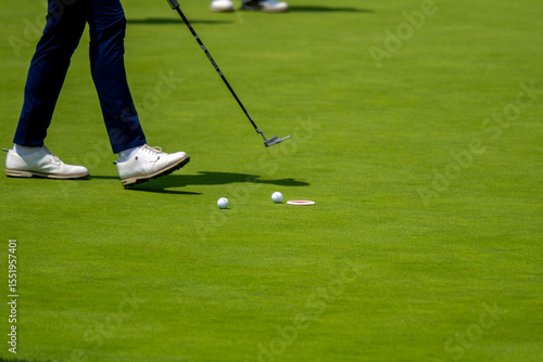 A golfer, mid-stride, walks past two golf balls and the hole on a vibrant green. The image captures a moment during a golf game, emphasizing movement and precision.