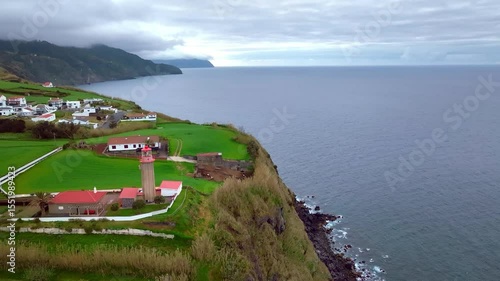 Aerial drone view of a lighthouse and a coastal village with red roofs and green fields on São Miguel Island, Azores, Portugal.