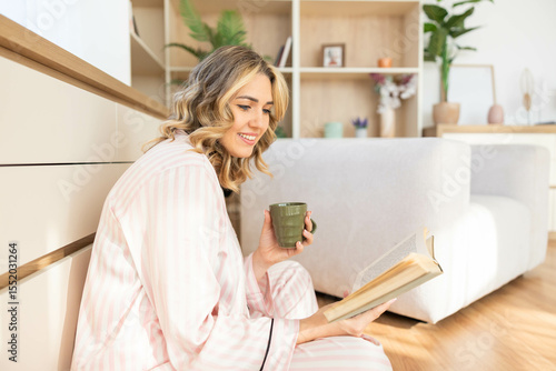 A relaxed woman reads on the floor with a cup of tea, surrounded by tranquility and the comforts of her inviting home.