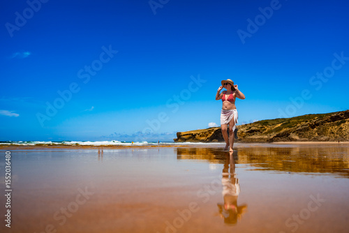 Beautiful mid adult woman model walking on sandy beach in summertime. Front view. Monte Clerigo beach on Algarve coast in Portugal