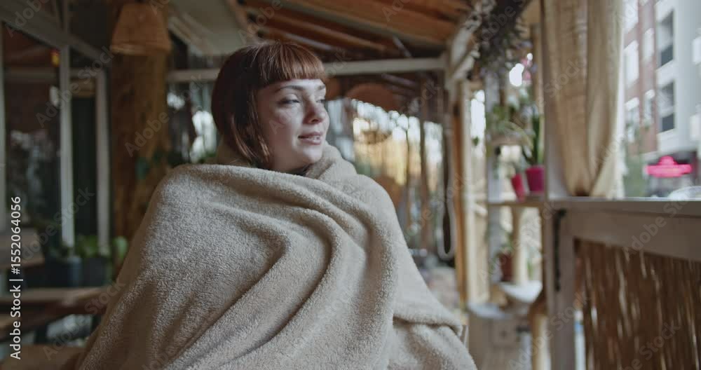 Young woman wrapped in cozy blanket, enjoying a peaceful moment on a cafe balcony, looking out at the city view. Young woman wrapped in cozy blanket, enjoying a peaceful moment on a cafe balcony, looking out at the city view.