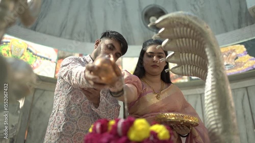 An Indian couple performs the sacred Abhishekam ritual to a Shiva Lingam in a Hindu temple. The woman is dressed in an elegant silk saree with jasmine flowers in her hair, and the man wears traditiona