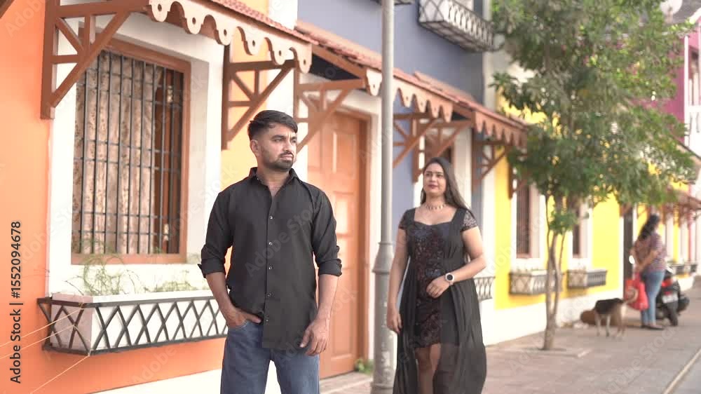A joyful Indian couple enjoying a candid moment while standing against a bright yellow wall. The woman is wearing a stylish black dress with sheer overlay, and the man is dressed casually in a black s A joyful Indian couple enjoying a candid moment while standing against a bright yellow wall. The woman is wearing a stylish black dress with sheer overlay, and the man is dressed casually in a black s