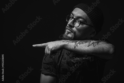 Moody black and white portrait of a tattooed male photographer holding a professional camera. Artistic low-key lighting highlights creative lifestyle concept