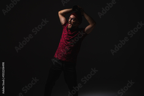 Stylish male photographer in red beanie and graphic tee posing in dramatic low key lighting, holding a camera against dark background. Urban creative vibe