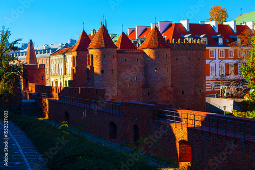 Historic brick fortifications with conical towers stand in the heart of Warsaw Old Town, bathed in warm sunlight. The medieval architecture contrasts beautifully with the colorful townhouses behind