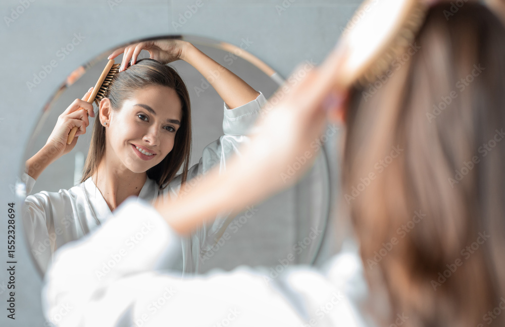 Naklejka premium Portrait Of Young Attractive Lady Combing Her Hair With Brush And Looking At Mirror While Standing Near Mirror In Bathroom, Beautiful Female Enjoying Self-Care, Selective Focus On Reflection