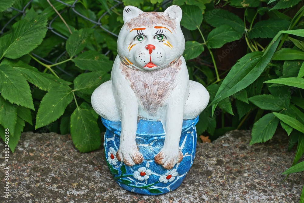 Fototapeta premium one large ceramic colored bank piggy bank with a figure of a cat stands on a gray table against the backdrop of green vegetation on a summer street