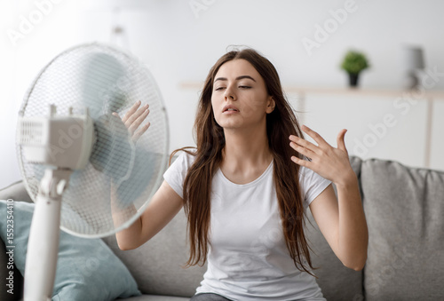 Sad young european woman suffers from unbearably too hot weather, catches cold air from fan in living room interior. Home without air conditioning, lady tired from summer heat, waves arms to cool down