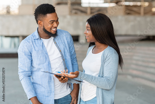 Black female volunteer conducting survey with young man on the street and writing down answers to clipboard, selective focus