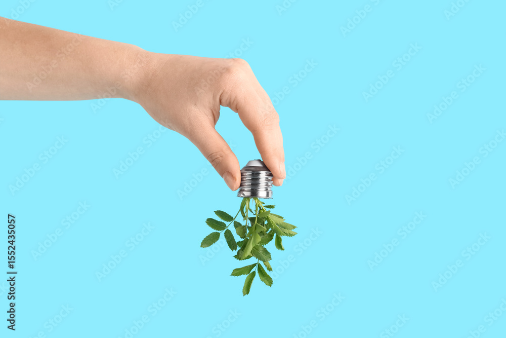 Fototapeta premium Female hand holding light bulb with plant leaves on blue background. Green technology concept