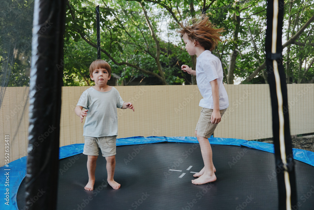 Obraz premium Joyful boys bouncing on a trampoline, enjoying childhood moments under a tree shade, exuding happiness and energy during playful summer days.