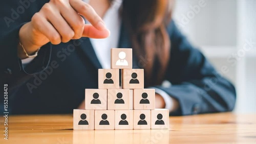 A person placing a wooden block with a person icon on top of a pyramid of similar blocks, suggesting a hierarchical organizational structure