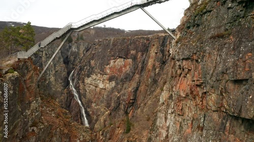 Voringsfossen waterfall gracefully cascades down vast rock face under bridge. Slender stream of water carves striking path through mountain landscape
