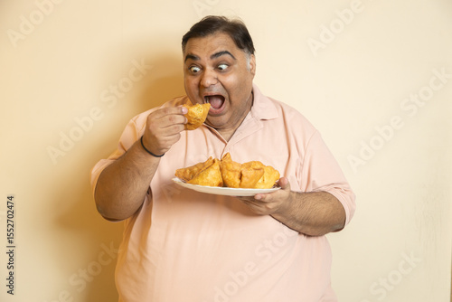 Hungary overweight fat indian man eating unhealthy samosa junk food. Chubby guy starving for fried snack. Plus size male crave for high calorie meal. Obesity and weight gain.