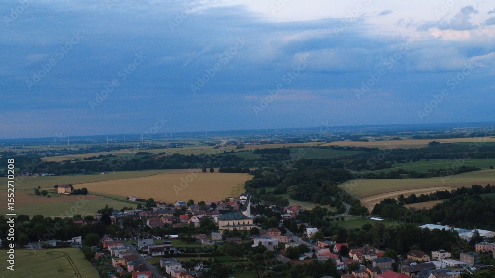 Fototapeta premium Aerial drone view of a peaceful rural landscape during summer sunset in Silesia, Czech Republic. The horizon glows with warm colors above rolling fields, small village houses, and distant Jesenik.