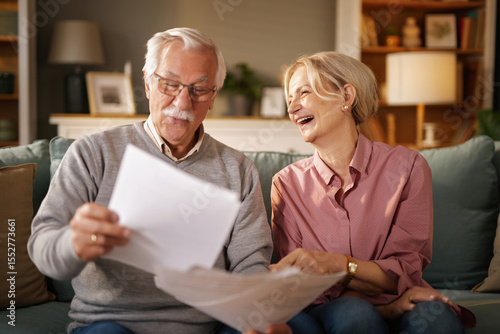 Картината върху платно A senior man with glasses reads papers while a smiling senior woman sits beside him on the sofa in their home