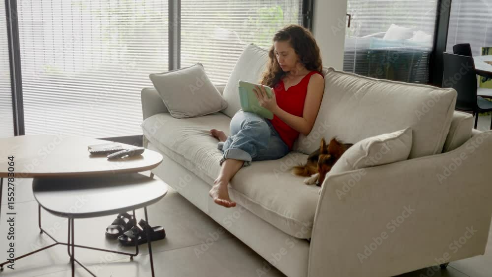 Woman sitting on couch working on digital tablet device and pen in living room. Focused and concentrated female student or freelancer working comfortably from home with her pet dog by her side Woman sitting on couch working on digital tablet device and pen in living room. Focused and concentrated female student or freelancer working comfortably from home with her pet dog by her side